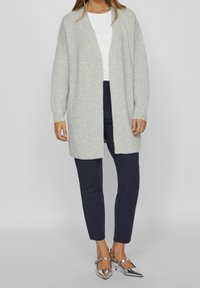Světlý šedý kardigan s žebrovanou texturou, otevřený vpředu a s dlouhými rukávy, kombinovaný s tmavě námořnickými kalhotami a stříbrnými botami.