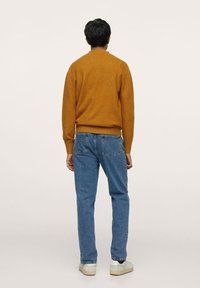 Musztardowy wełniany sweter z prążkowanym dołem, w zestawieniu z niebieskimi dżinsami. Osoba nosi białe trampki, widok z tyłu.