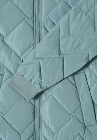Giacca trapuntata azzurro chiaro con chiusura a zip, polsini a costine e un motivo a zigzag sul tessuto. Texture morbida e liscia.