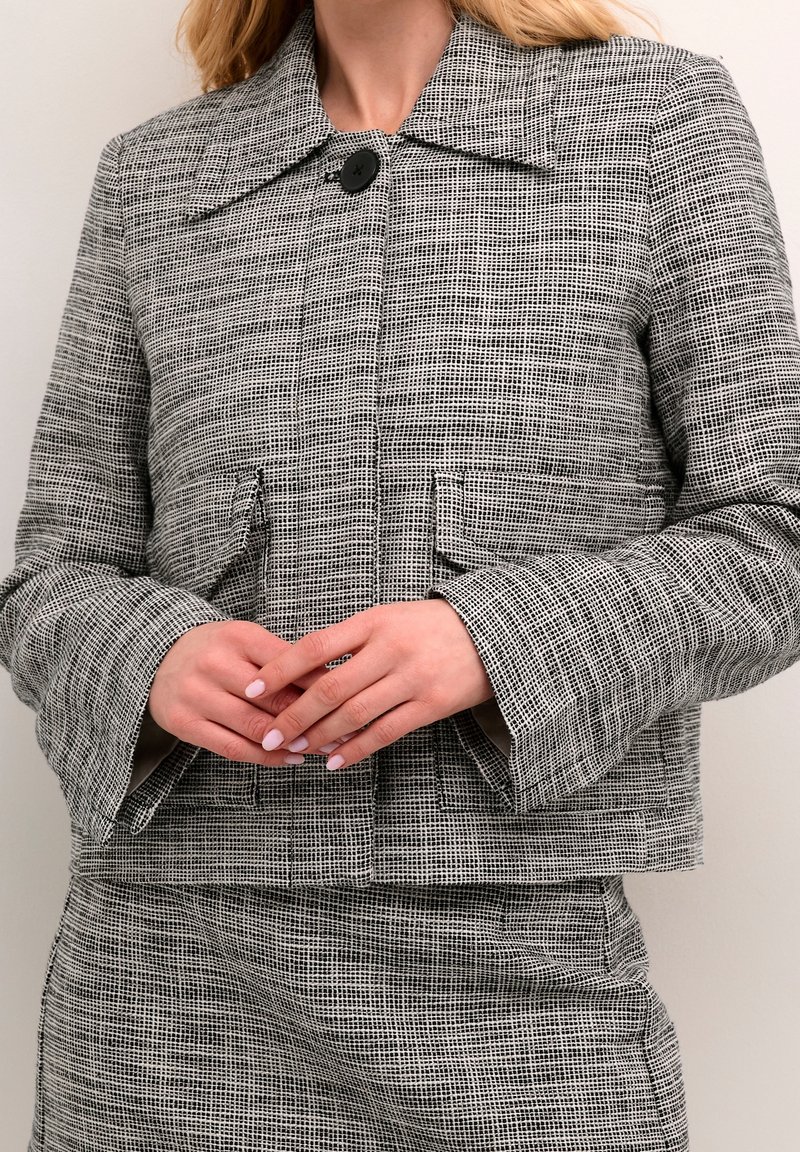 Femme portant une veste texturée gris et noir à motifs avec de grandes poches et une jupe assortie, les mains jointes à la taille.