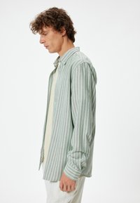 Camisa de botones con rayas verticales verdes y blancas, con cuello y mangas remangadas, llevada sobre una blusa de color claro.