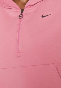 Personne portant un sweat à capuche Nike rose avec une fermeture éclair mi-longue et une poche frontale en kangourou, montrant le haut du torse et la zone du cou.