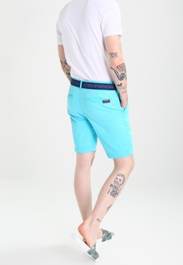 Homme portant un t-shirt blanc, un short turquoise, une ceinture bleu marine et des sandales, montrant des tatouages sur les bras et les jambes, debout sur un fond blanc.