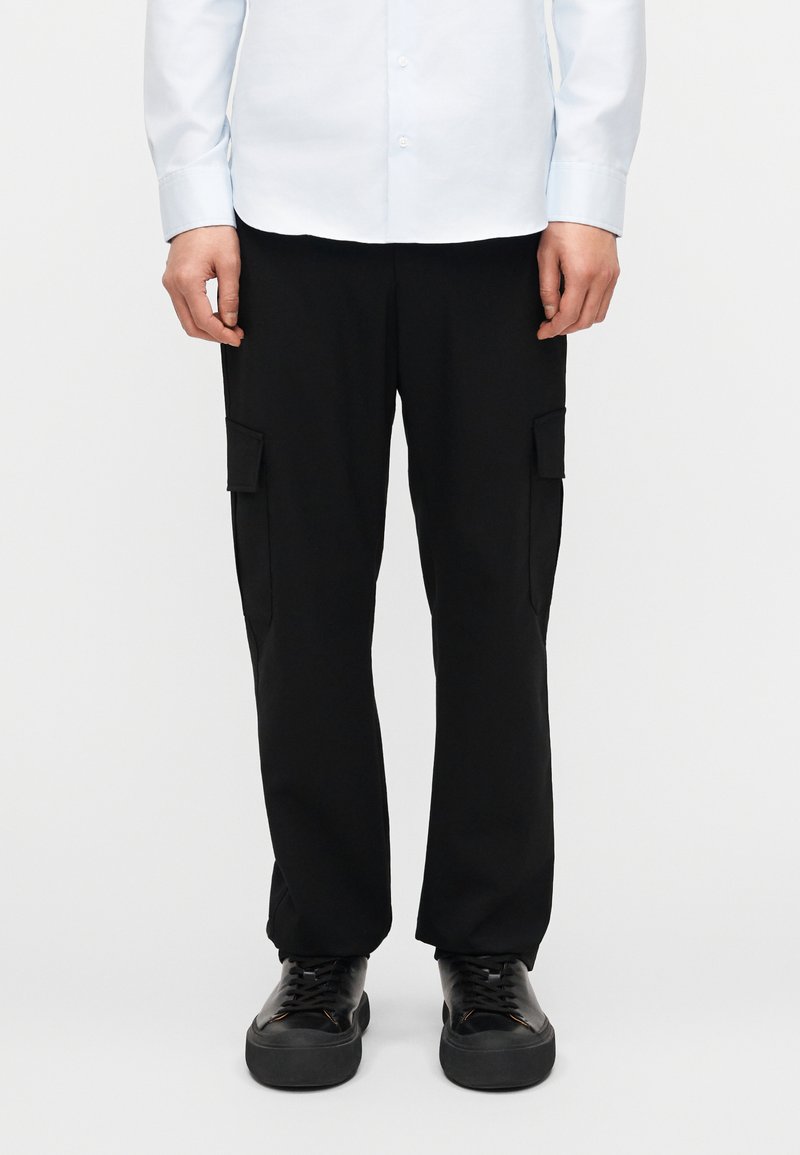 Άτομο που φοράει μαύρο παντελόνι cargo με πλαϊνές τσέπες, μαύρα αθλητικά παπούτσια και ανοιχτό μπλε πουκάμισο με κουμπιά, σε απλό φόντο.