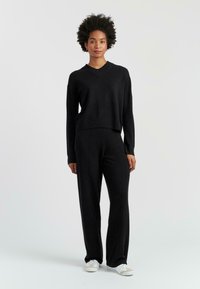 Pull en V noir en tricot avec des détails côtelés, associé à un pantalon noir à jambes larges. Modèle portant des baskets blanches, sur un fond neutre.