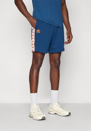 Uomo che indossa pantaloncini blu Kappa con logo arancione e strisce laterali, calzini bianchi e sneakers beige, in piedi contro uno sfondo semplice.