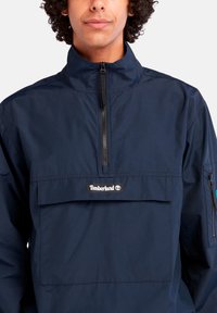Jaunas žmogus vilkintis tamsiai mėlyną pusiau užtraukiamą striukę su priekyje esančia kišene ir Timberland logotipu ant krūtinės, priešais baltą foną.