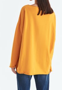 Ylimittainen villapaita kirkkaan oranssina, tehty pehmeästä kankaasta, jossa leveä, venepääntie ja laskokset hartioiden kohdalla. Helma kaartuu hieman.