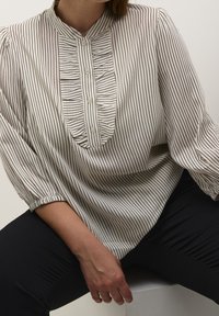 Blouse crème avec des rayures noires verticales, col mandarin et patte froncée. Présente des fermetures à boutons et des manches bouffantes froncées.