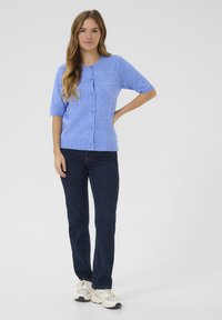 Világoskék, rövid ujjú kardigán, kerek nyakkivágással, elől gombos záródással és bordázott szegéllyel, sötét farmernadrág és világos edzőcipő felett viselve.