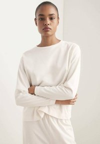 Chemise longue à manches longues beige clair, col rond, coupe décontractée, en tissu doux, avec un détail torsadé à l'ourlet et une texture lisse dans son ensemble.
