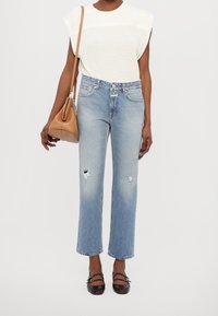 Jean taille haute bleu clair avec détails usés, associé à un haut sans manches blanc texturé et un sac à épaule marron clair. Des ballerines noires complètent le look.