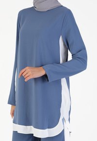 Tunica blu realizzata in tessuto leggero. Presenta maniche lunghe, spacchi laterali e una vistosa rifinitura bianca. Vestibilità morbida e rilassata.