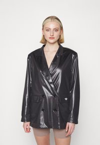 Must mustriga mustriga, läikivast kangast, kahekordse nööbiga bleiser, millel on kaks esilocust ja väike valge logo vasakul.