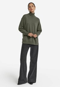 Grüner Rollkragenpullover aus weichem Material, mit lässigem Schnitt, gerippten Akzenten und langen Ärmeln, kombiniert mit dunklen, weiten Jeans.