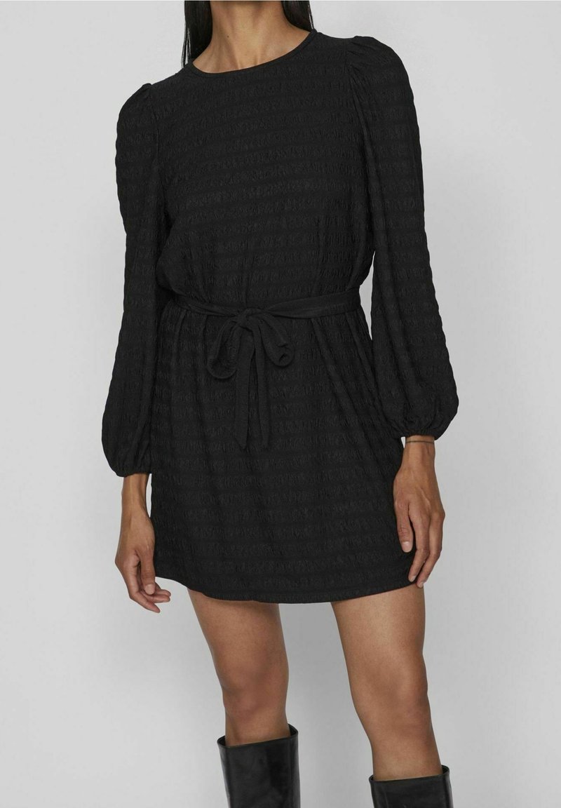 Femme portant une mini-robe noire texturée à manches longues avec des épaules bouffantes et une ceinture nouée à la taille, associée à des bottes noires montantes.