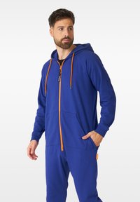 Blue hoodie jumpsuit made of cotton. Features an orange zipper, matching drawstrings, and front pockets. Smooth texture and relaxed fit.
