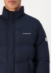Marineblauwe gewatteerde jas met hoge kraag, voorzien van het "GUESS JEANS" logo in het wit en een klein merkaccent op de mouw.