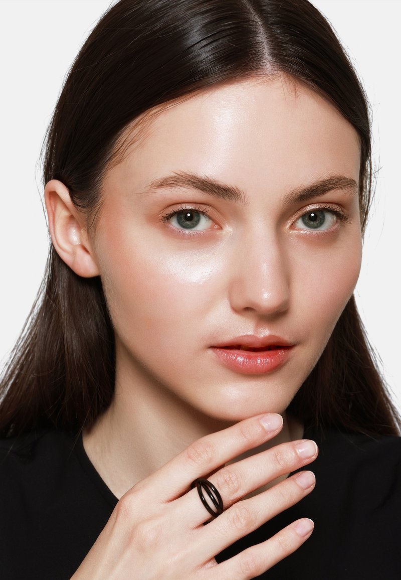 Zwarte metalen ring met een verweven ontwerp, model houdt deze tegen haar gezicht, gladde huid, natuurlijke make-up en lang donker haar, grijze ogen.