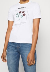 Hvid bomulds t-shirt med korte ærmer, der pryder et blomstermotiv i rød og grøn. Tekst trykt i sort. Båret med blå jeans.