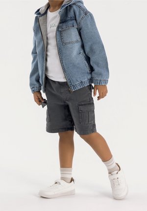 Spijkerjack met een grijze capuchon, wit T-shirt, grijze cargoshorts met zakken en witte sneakers met lichtgekleurde sokken.