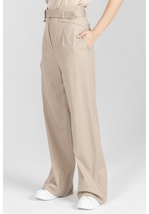 Beżowe, szerokie spodnie z wysokim stanem i dopasowanym paskiem, noszone z białymi trampkami, ręka modela w kieszeni.
