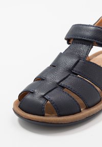 Ciemnogranatowy skórzany sandał z plecionym wzorem, odkrytymi palcami i regulowanym paskiem. Podeszwa zewnętrzna ma naturalne, teksturowane wykończenie.