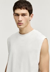 Weißes ärmelloses Shirt aus weichem Stoff, mit rohen Kanten und entspannter Passform, das eine glatte Textur und ein minimales Design zeigt.