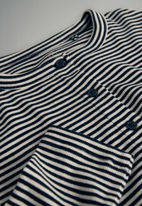 Camisa de rayas con franjas horizontales en azul marino y crema, tres botones negros y un pequeño bolsillo en el lado izquierdo. Textura suave y cuello acanalado.