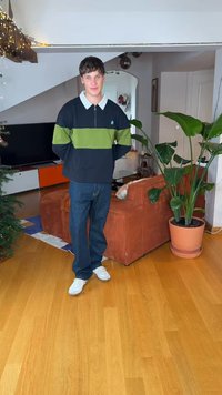 Jauns vīrietis stāv viesistabā ar koka grīdu, oranžu dīvānu, lielu podu augu, Ziemassvētku eglīti un TV skapīti ar oranžām un baltām atvilktnēm.