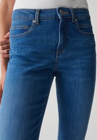 Jeans en denim bleu avec un design ajusté, présentant un style classique à cinq poches, des détails en métal argenté et des surpiqûres contrastantes.
