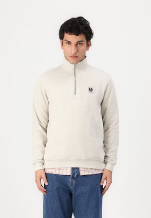 Sudadera con cremallera de color beige claro, con cuello alto, puños y dobladillo de canalé; presenta un pequeño parche de logo en el pecho. Lleva encima una camisa de cuadros.