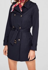 Granatowy płaszcz trenchowy z podwójnym zapięciem, złotymi guzikami, paskiem w talii oraz pagonami na ramionach. Zawiera czerwoną kwiecistą chustę na szyi.