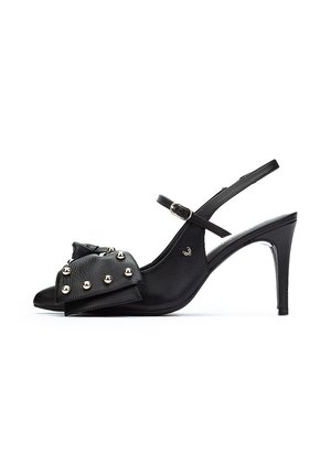 Zapatos de tacón negro con tiras, punta afilada, que presentan un gran lazo y tachuelas doradas en la parte delantera. Textura de cuero suave; tacón de aguja delgado.