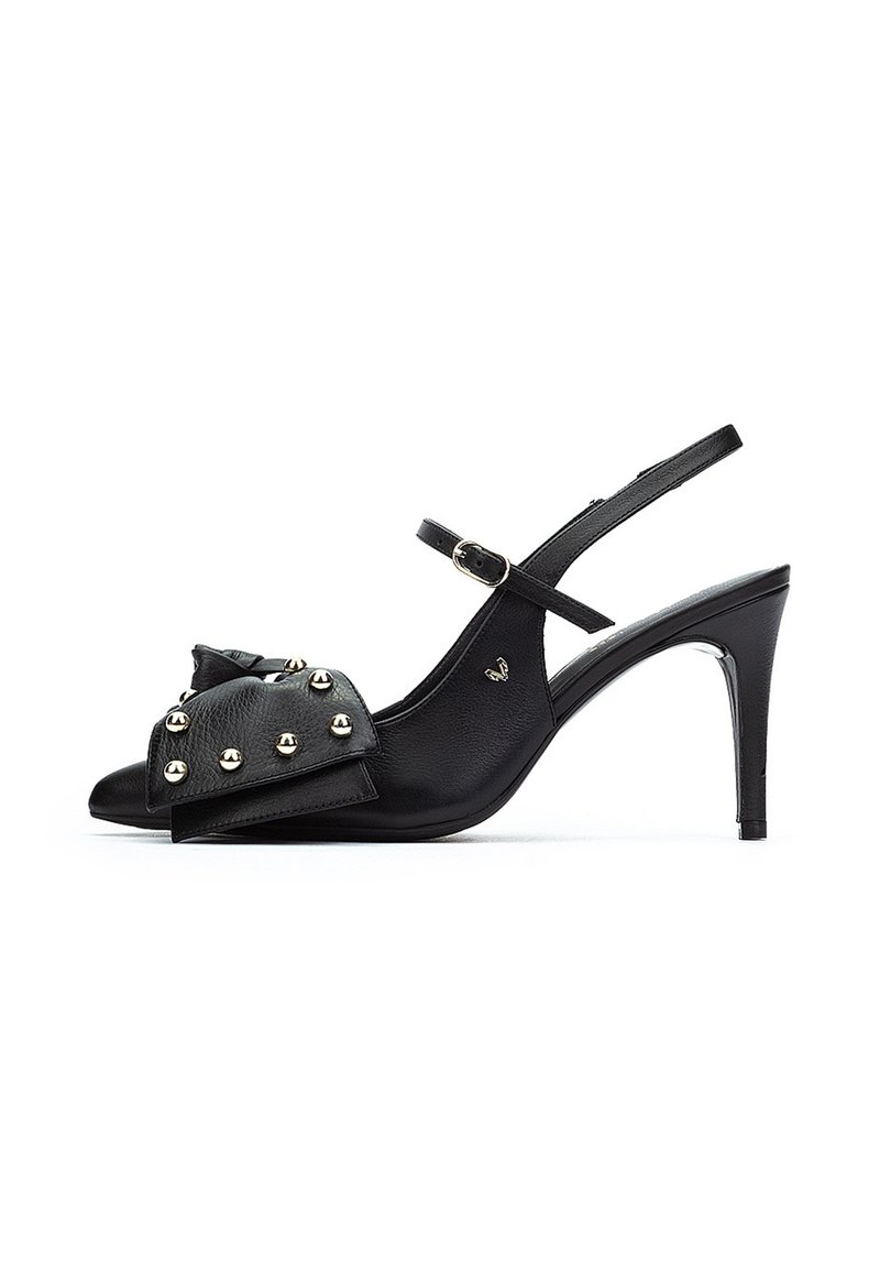 Zapatos de tacón negro con tiras, punta afilada, que presentan un gran lazo y tachuelas doradas en la parte delantera. Textura de cuero suave; tacón de aguja delgado.