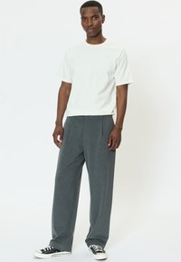 Biele tričko s krátkym rukávom v kombinácii s voľnými tmavosivými nohavicami. Nohavice majú záhyby a sú nosené s čierno-bielymi teniskami.