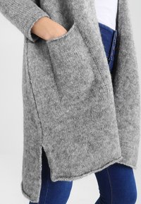 Harmaa neulottu neuletakki, jossa on pitkät hihat, sivutaskut ja hieman pidempi takapani, päällä sinisiä farkkuja. Pehmeä, tekstuuriensa ansiosta mukava kangas.