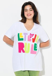 T-shirt blanc avec un col en V, arborant des lettres en blocs audacieuses et multicolores écrivant "LAISSE L'AMOUR RÉGNER" en vert, jaune, rose, violet et rouge.