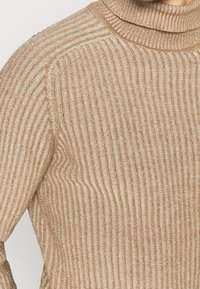 Beiger, gerippter Strickpullover mit hohem, rundem Ausschnitt und vertikalen Streifen, die seine weiche Textur und seine figurbetonte Form betonen.