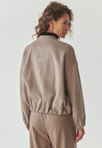Světlě hnědý oversized svetr s žebrovaným černým límcem a elastickým lemem. Měkká, texturovaná látka; spadlé ramena a dlouhé rukávy.
