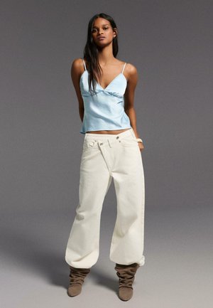 Vrouw met lichtblauw spaghettihemd, oversized off-white broek en bruine loszittende laarzen, staand tegen een grijze achtergrond.