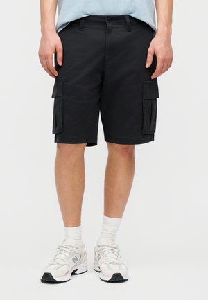 Πόδια που φορούν μαύρα cargo σορτς με πλαϊνές τσέπες, λευκές κάλτσες και λευκά αθλητικά παπούτσια New Balance, μπροστά σε απλό ανοιχτόχρωμο φόντο.