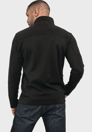 Schwarzer Pullover mit hohem Kragen, gerippter Textur und langen Ärmeln. Verfügt über einfache Nähte und einen geraden Saum, getragen über dunklen Jeans.
