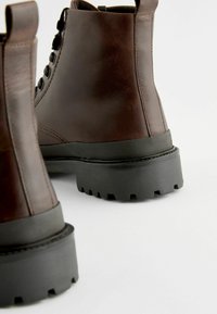 Botas de cuero marrón hasta el tobillo con una suela de goma negra texturizada y cordones, que cuentan con punta redondeada y una lengüeta en la parte posterior para facilitar su uso.