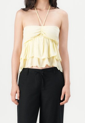 Femme portant un crop top halter à couches jaune pâle associé à un pantalon noir taille haute avec cordon de serrage sur un fond uni.