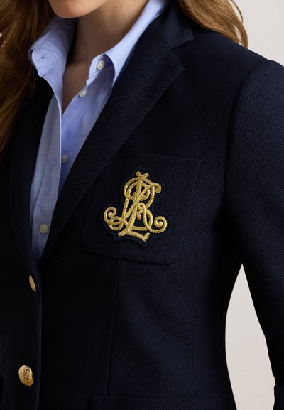 Gros plan sur une personne portant un blazer bleu marine avec un monogramme brodé en or sur la poche poitrine et des boutons dorés, par-dessus une chemise bleu clair boutonnée.