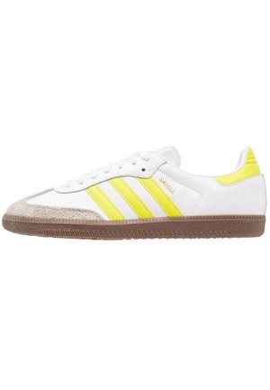 Baskets blanches Adidas Samba avec des bandes jaunes, embout en daim beige et semelle marron texturée, orientées vers la droite sur fond blanc.