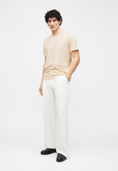 Άνδρας με σγουρά μαλλιά, φορώντας μπεζ κοντομάνικο πουκάμισο, λευκό παντελόνι με φαρδιά πόδια και μαύρα παπούτσια, στέκεται με το ένα χέρι στην τσέπη.