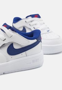 White leather sneakers with blue accents, featuring a distinctive Nike swoosh, textured sole, and white laces. Extra padding at the collar.