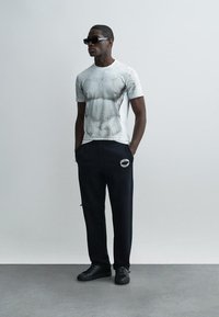 Homme portant des lunettes de soleil noires, un T-shirt blanc à motifs, un pantalon de survêtement Moschino noir et des baskets noires, debout les mains dans les poches.